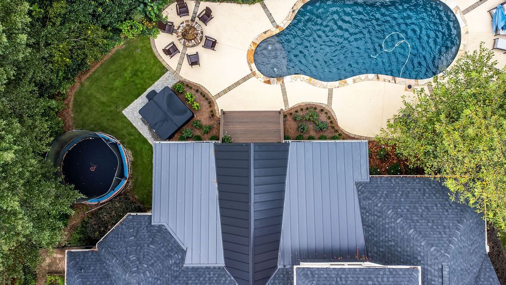Aerial view of a backyard with a swimming pool, patio seating, deck, and house roof.