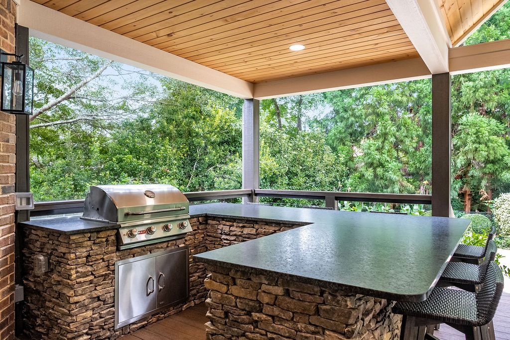 Covered outdoor kitchen with stone countertops, grill, and bar seating overlooking greenery