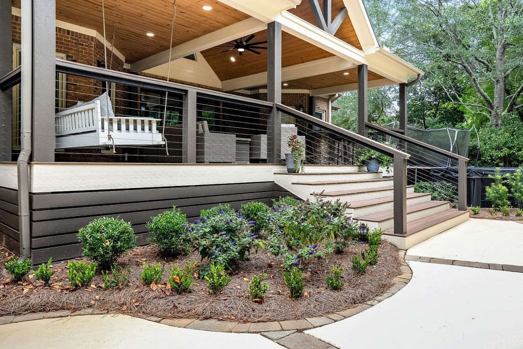 Covered porch with white swing, wooden steps, and landscaped flower beds beside a sidewalk