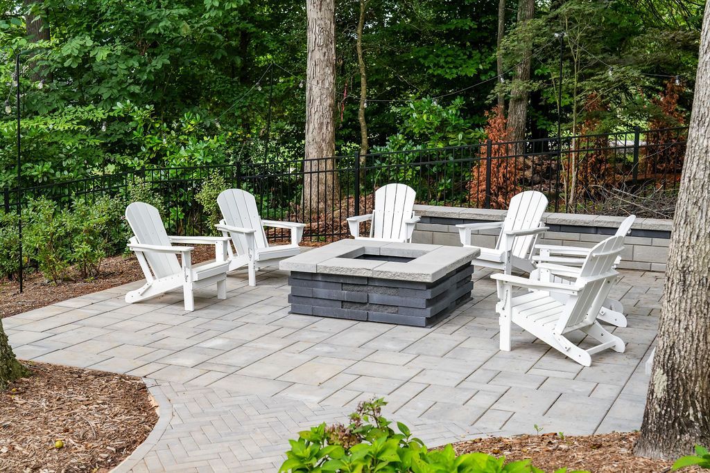 Outdoor patio with white Adirondack chairs вокруг a stone fire pit, surrounded by trees and gravel