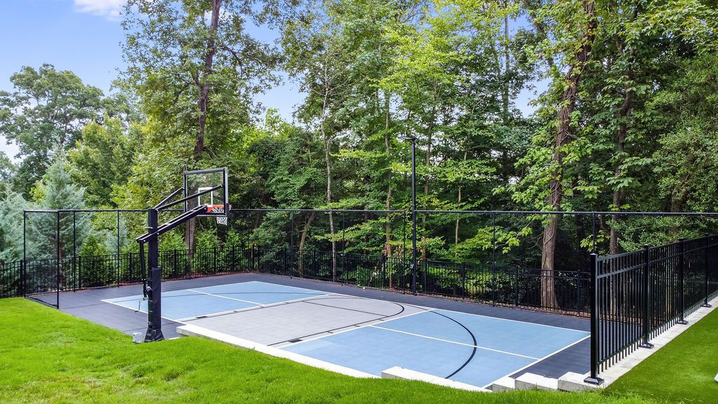Outdoor basketball court with black fence, blue-and-gray surface, and trees surrounding it