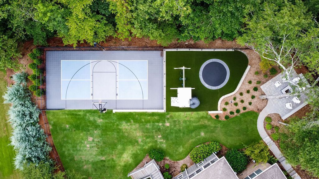 Aerial view of a backyard with a basketball court, trampoline, lawn, and surrounding trees.