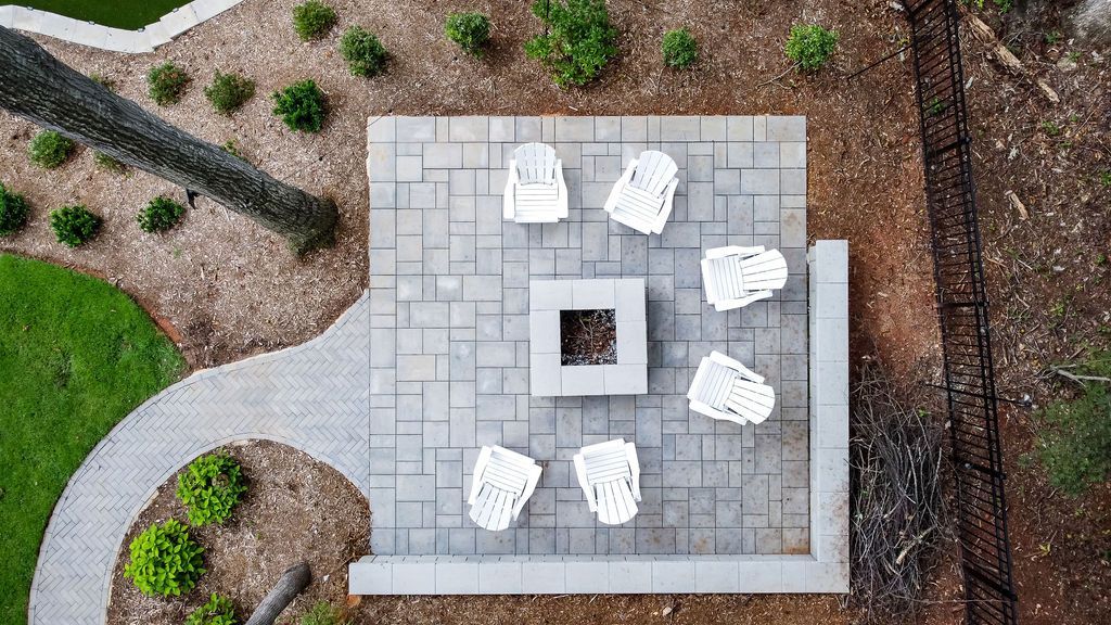 Overhead view of a gray patio with white chairs around a square fire pit, beside a curving path and landscaping