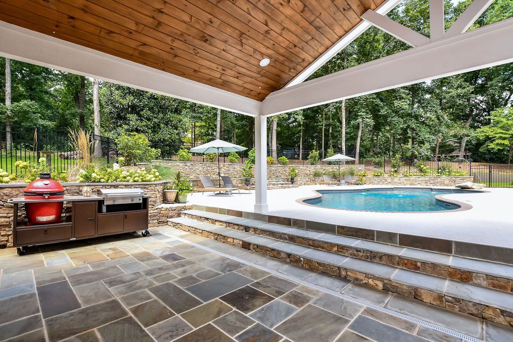 Covered patio with bluestone, outdoor kitchen, stone steps, and a pool overlooking a wooded yard