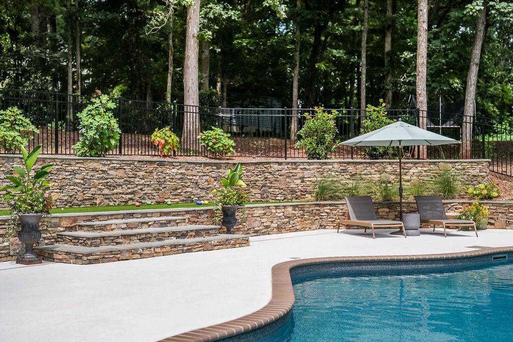 Backyard pool with stone patio, lounge chairs, umbrella, and lush trees in the background