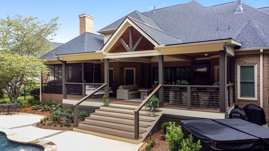 Back porch of a rustic house with stairs, cable railing, columns, and a gray roof under a sunny sky