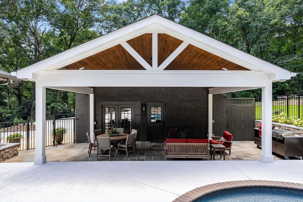 Covered patio with white gabled roof, seating area, red chaise, and pool edge in front of a house