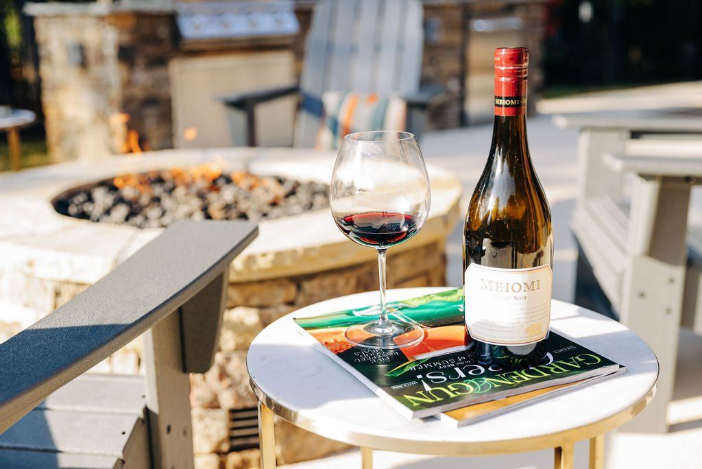 Wine bottle and glass on an outdoor table beside a fire pit, with a menu in sunlight.