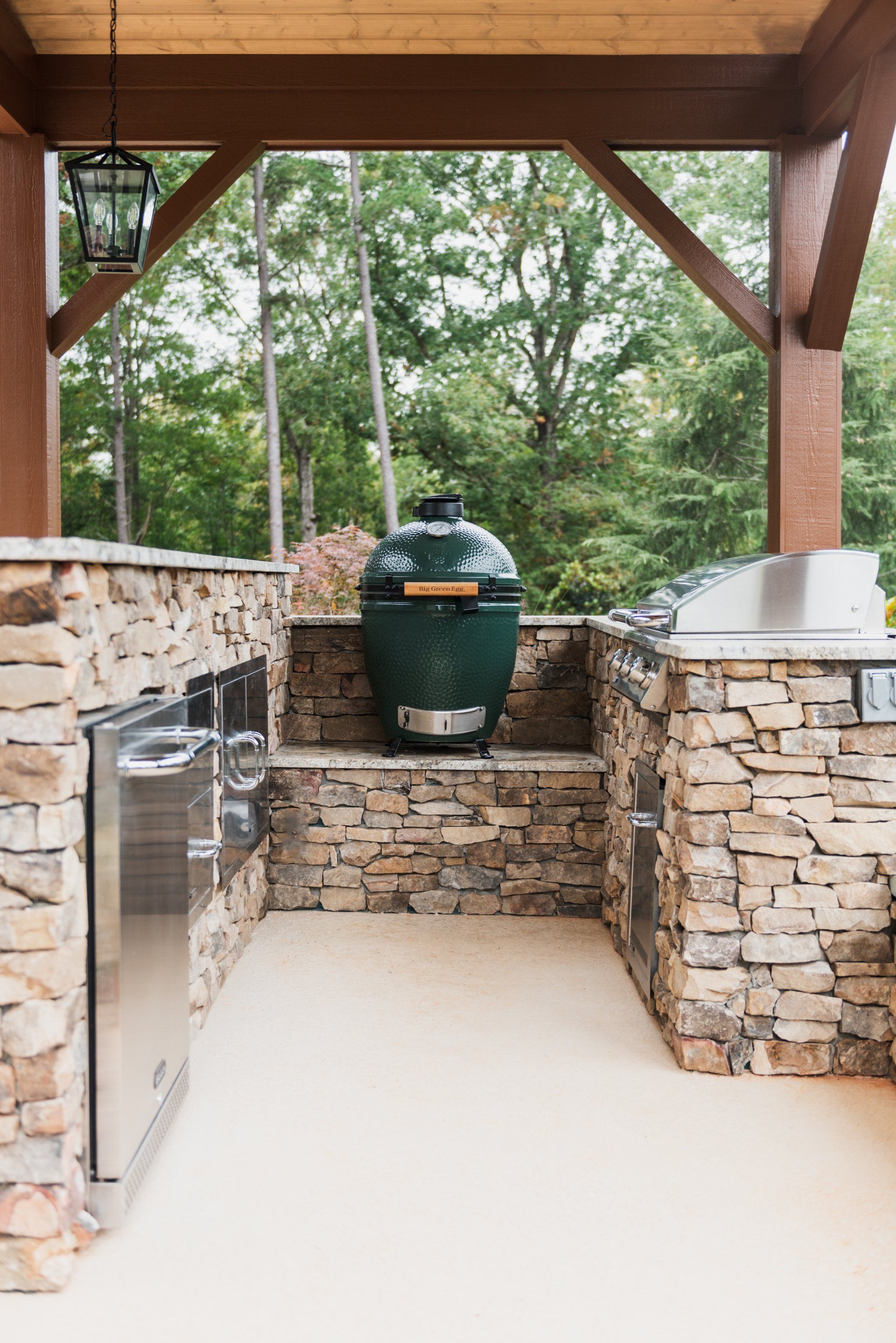 Covered outdoor kitchen with stone grill island and green barbecue smoker under a wooden pavilion.