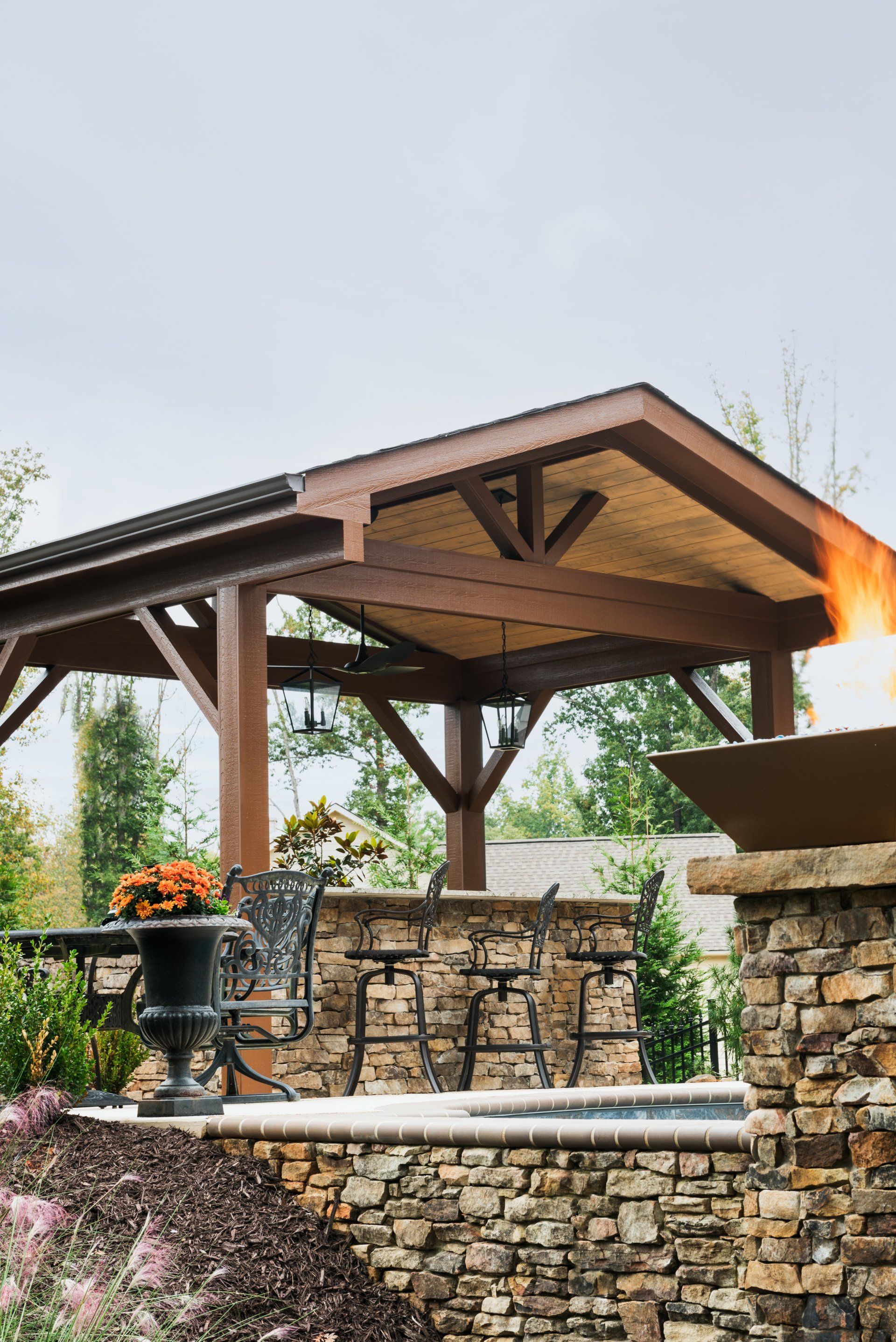 Stone outdoor fireplace beneath a wooden pavilion with potted flowers and stacked stone seating