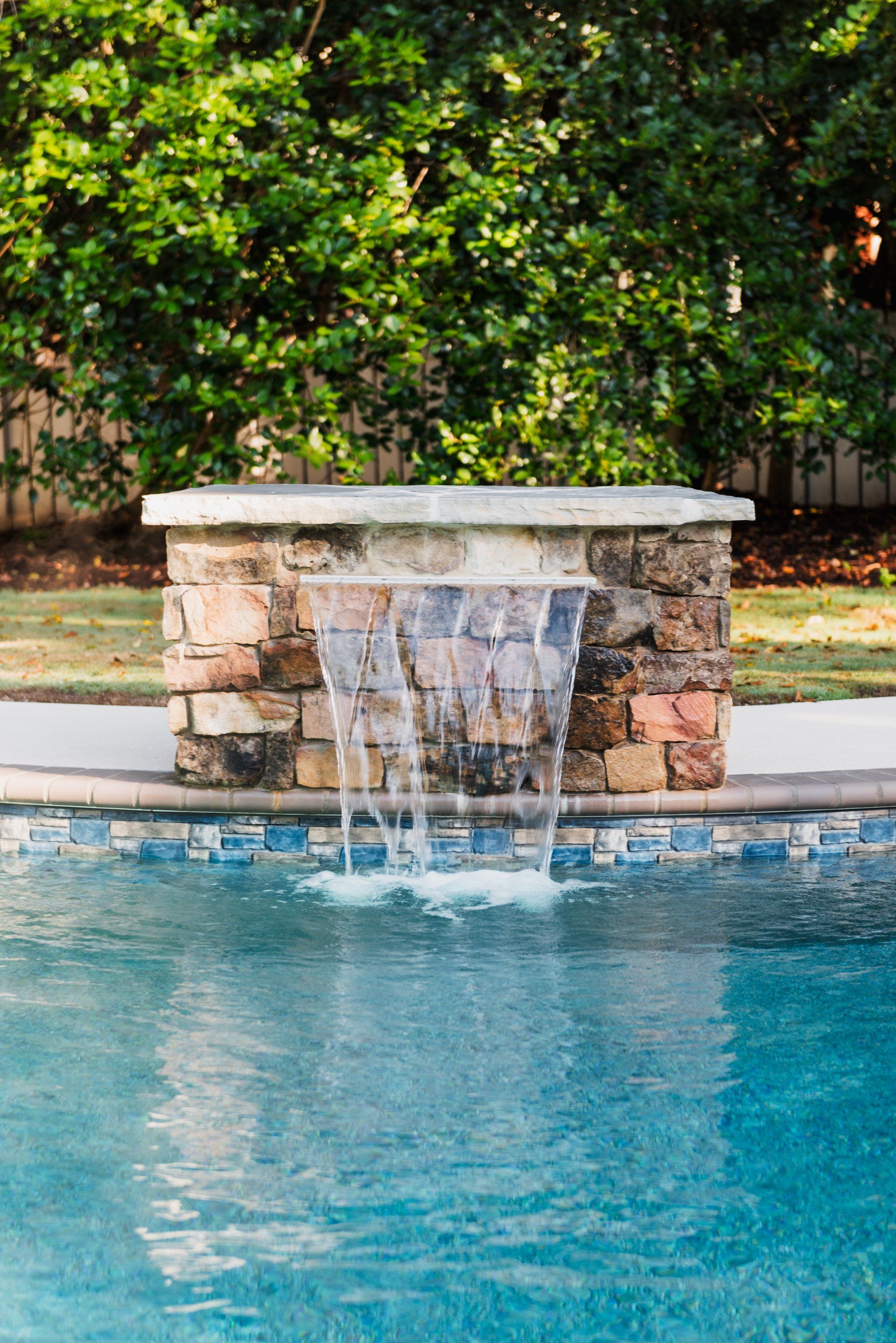 Stone waterfall fountain pouring into a blue pool with lush greenery behind it