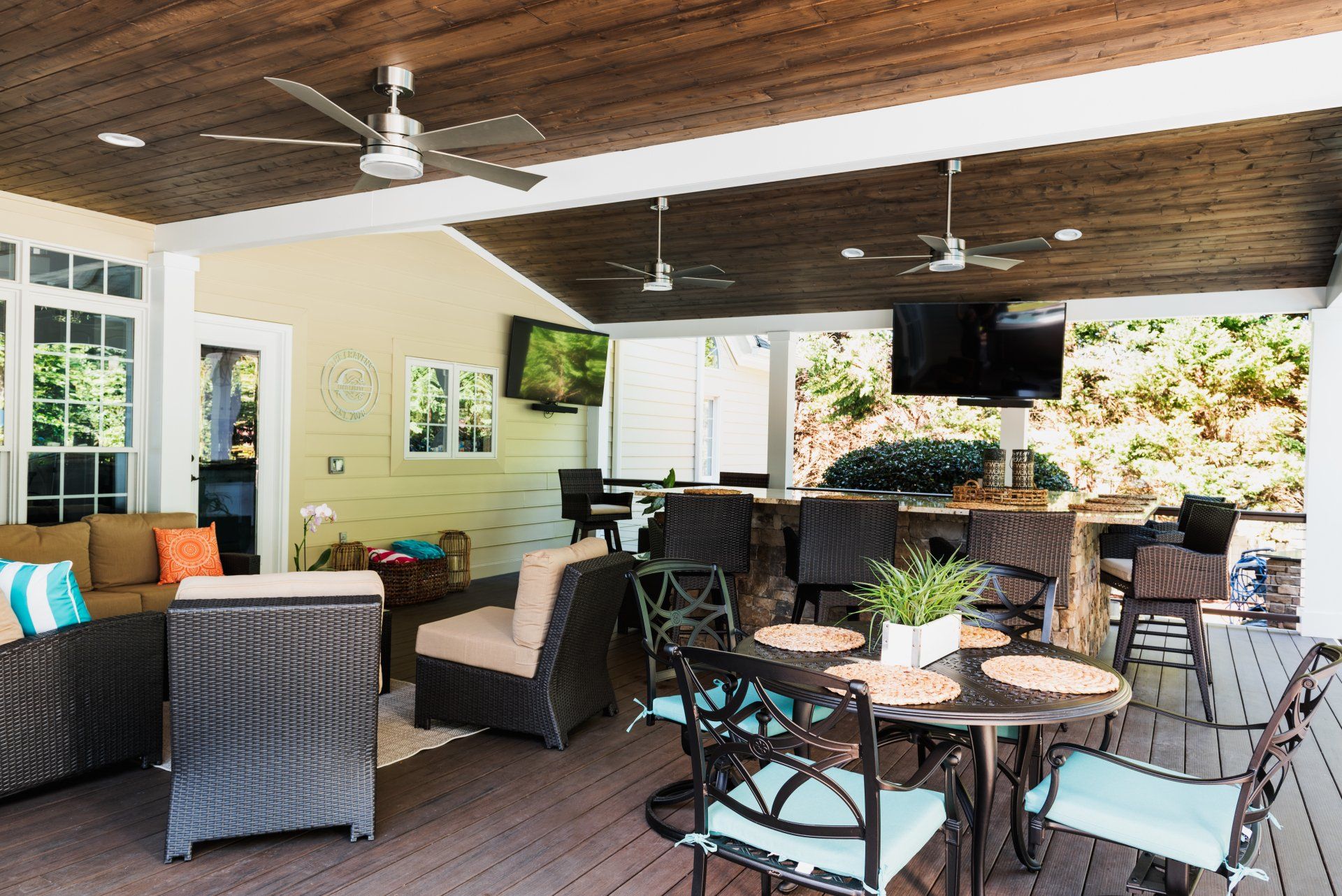 Covered patio with wicker seating, ceiling fans, and outdoor TV, overlooking greenery
