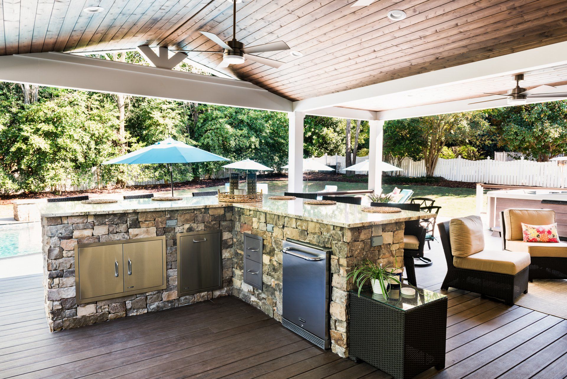 Covered outdoor poolside bar with stone counter, grill, stools, and lounge seating under a wood ceiling.