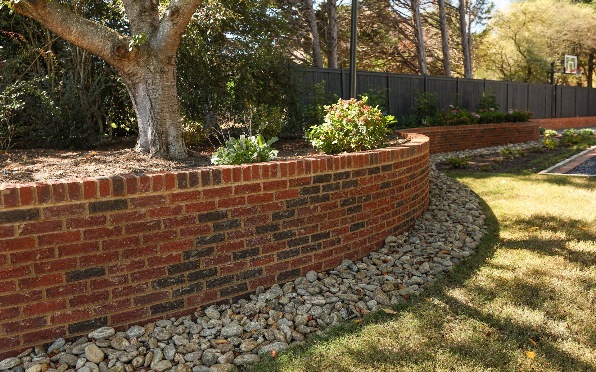 Curved red brick retaining wall edging a landscaped yard with rocks, grass, and trees in sunlight