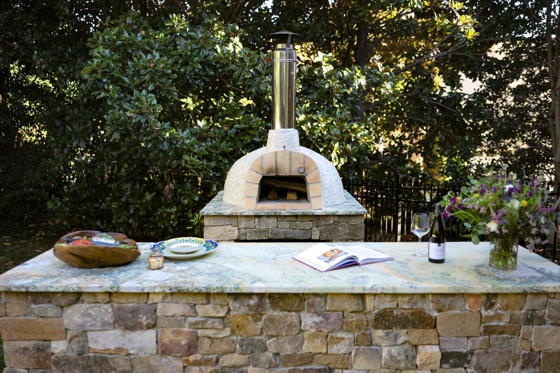 Outdoor stone pizza oven on a patio table with serving items and flowers, set against leafy greenery