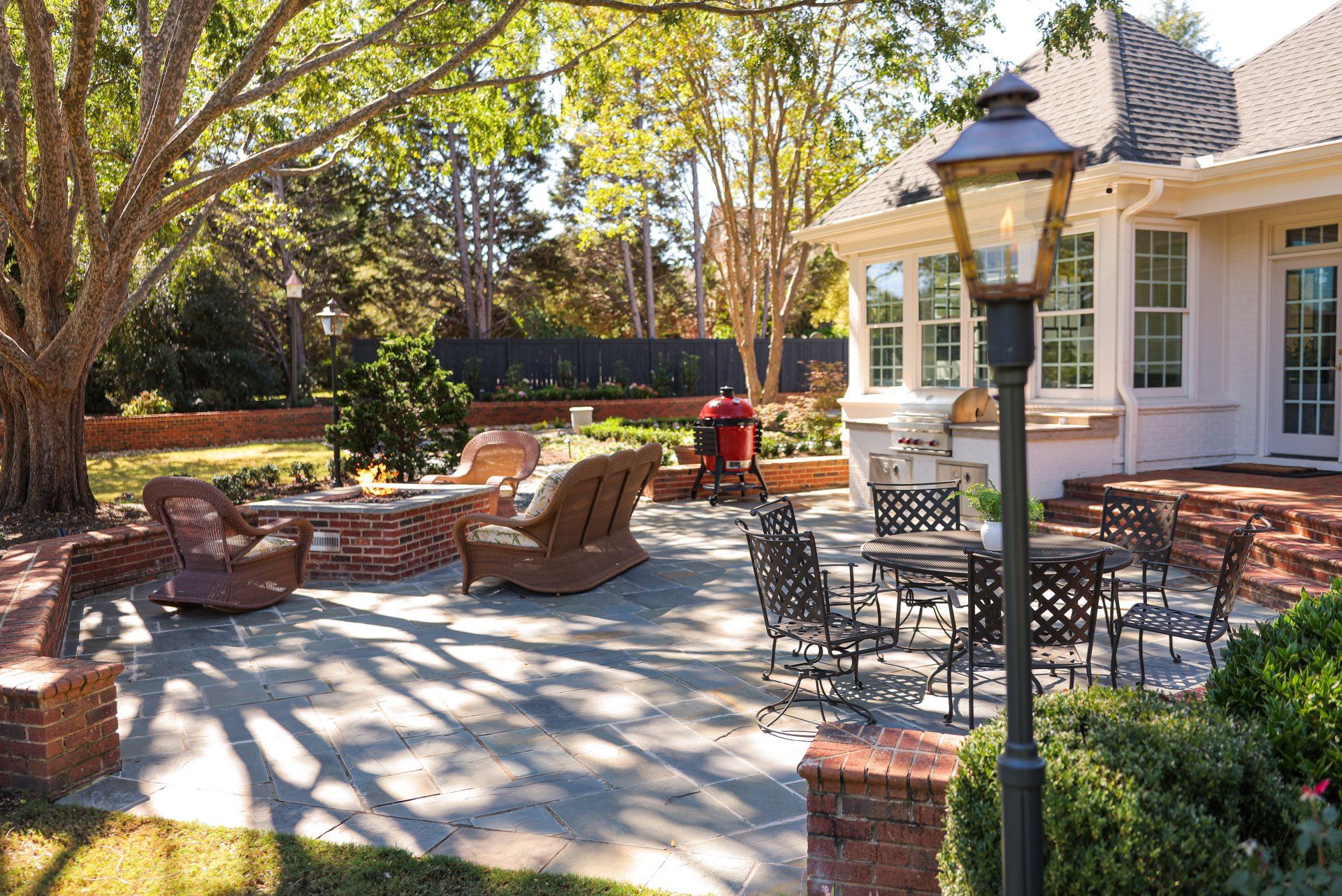 Brick patio with wicker chairs and tables under trees beside a house, lit by sunlight