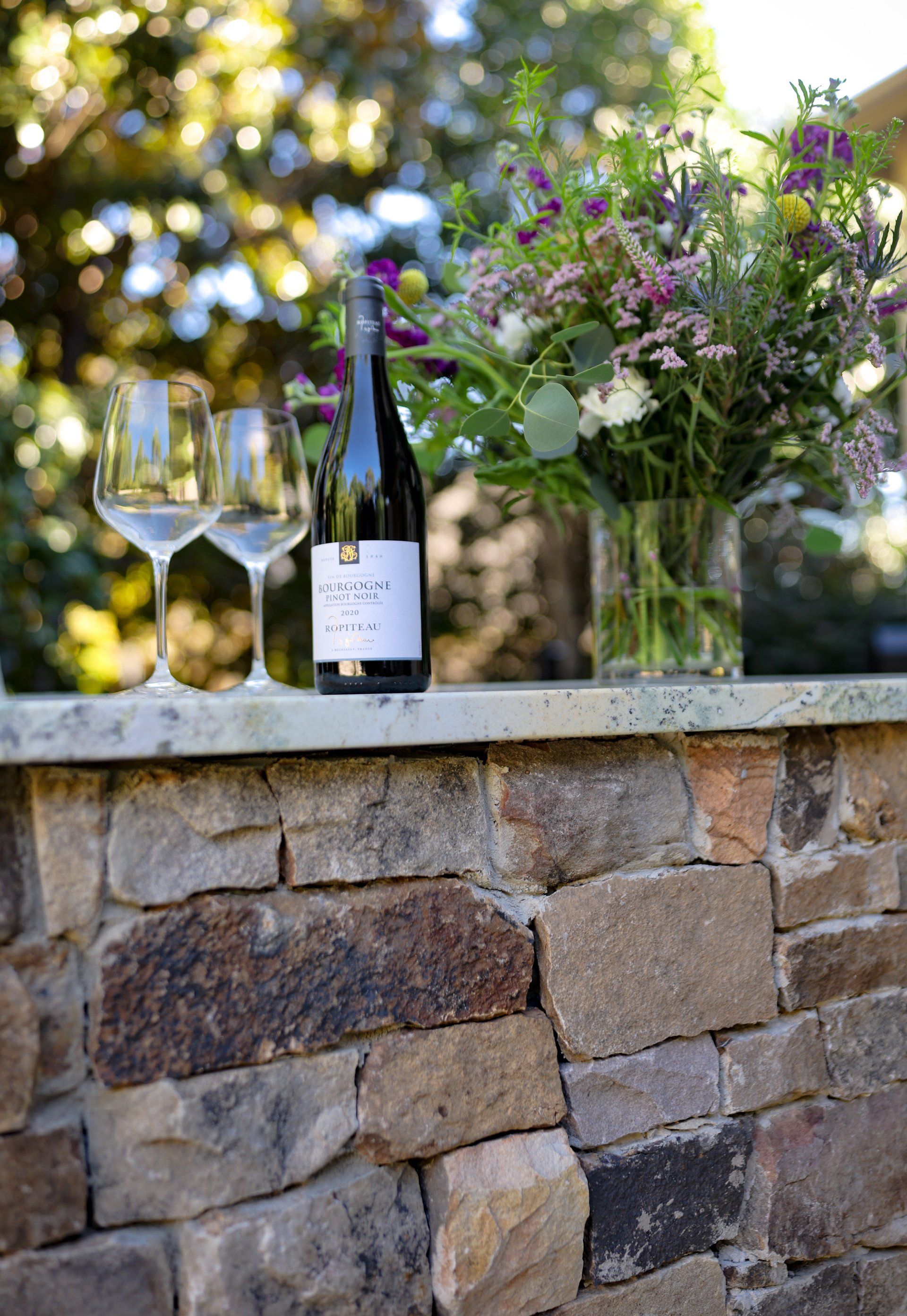 Wine bottle and two glasses on a stone ledge beside a colorful flower arrangement outdoors