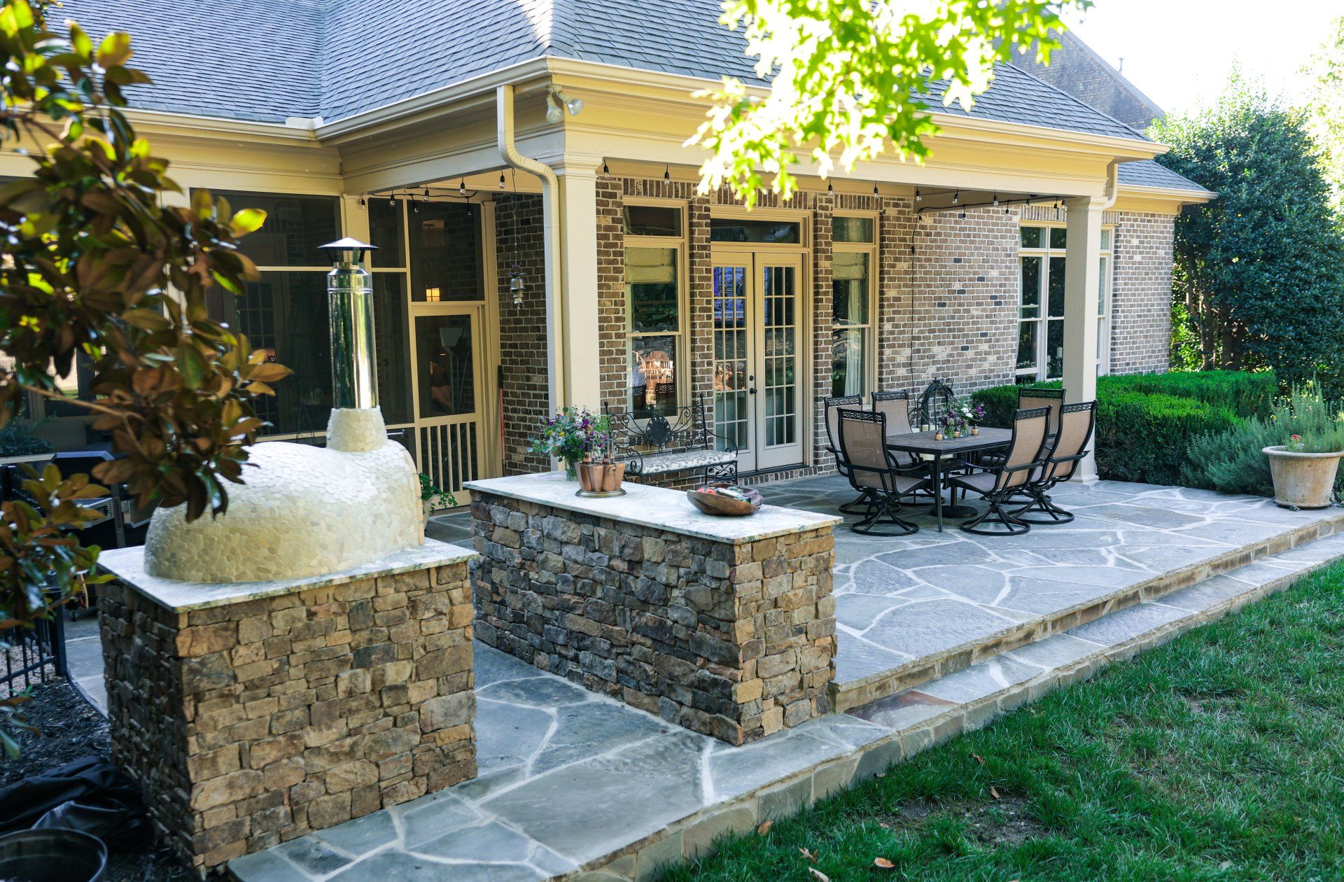 Patio with stone fountain, outdoor dining table, and chairs beside a beige house under leafy trees