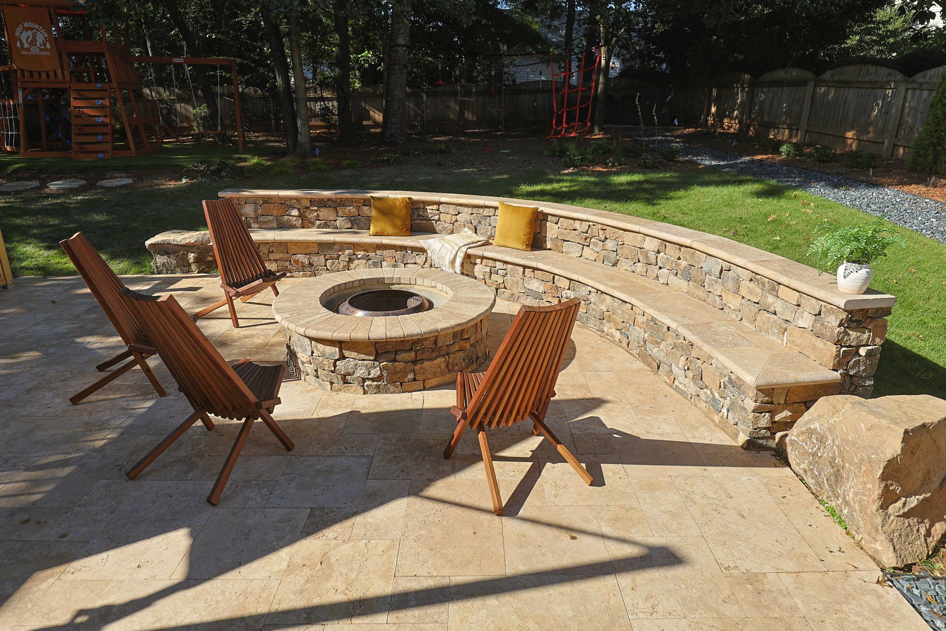 Stone fire pit with curved bench seating and wooden chairs in a sunny outdoor patio area