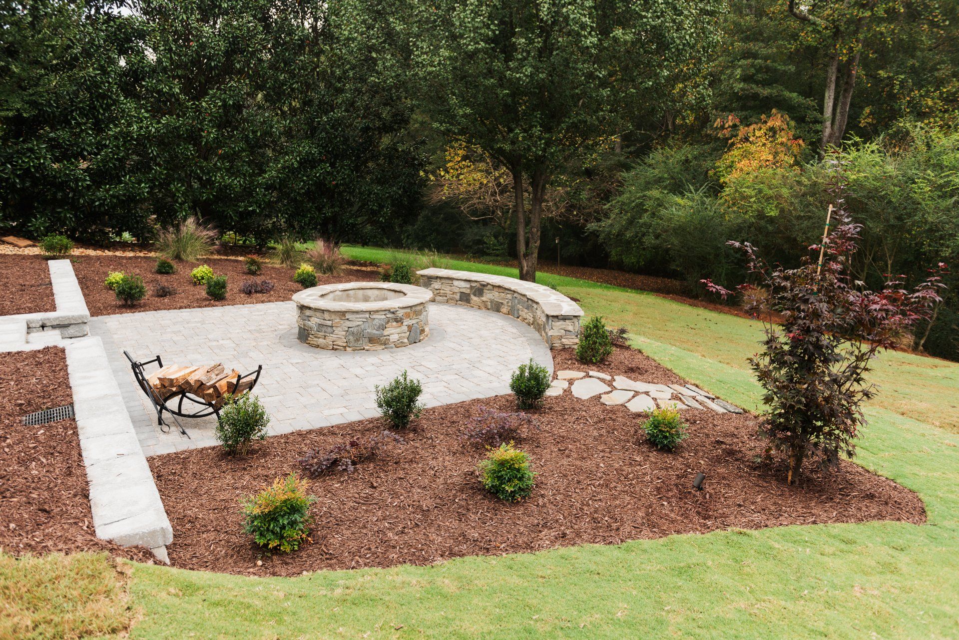 Landscaped garden with stone fire pit, mulch beds, and small shrubs surrounded by grass and trees