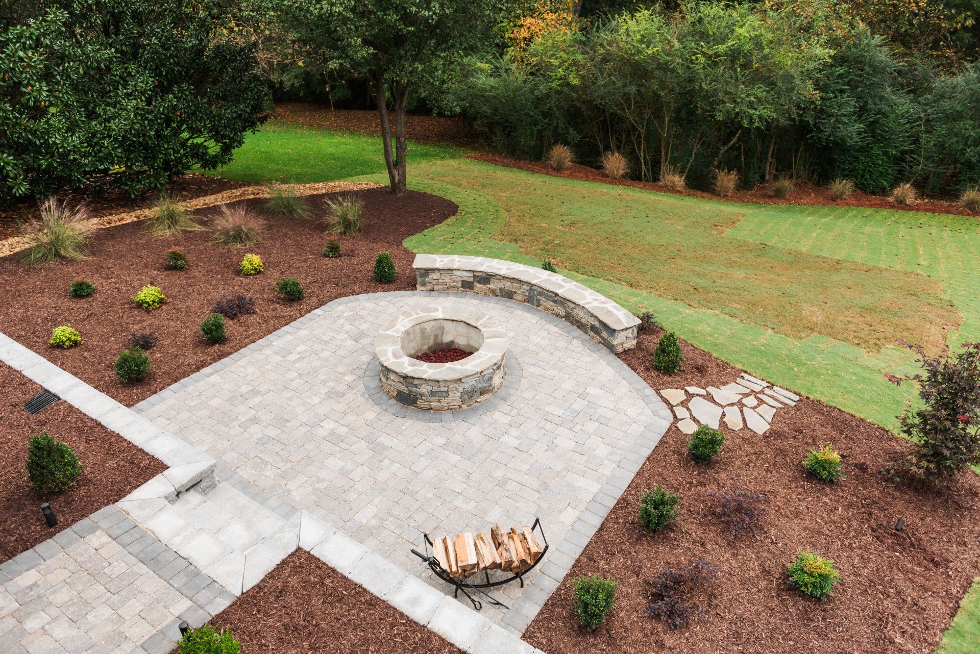 Backyard stone fire pit patio with curved seating wall, surrounded by mulch and shrubs