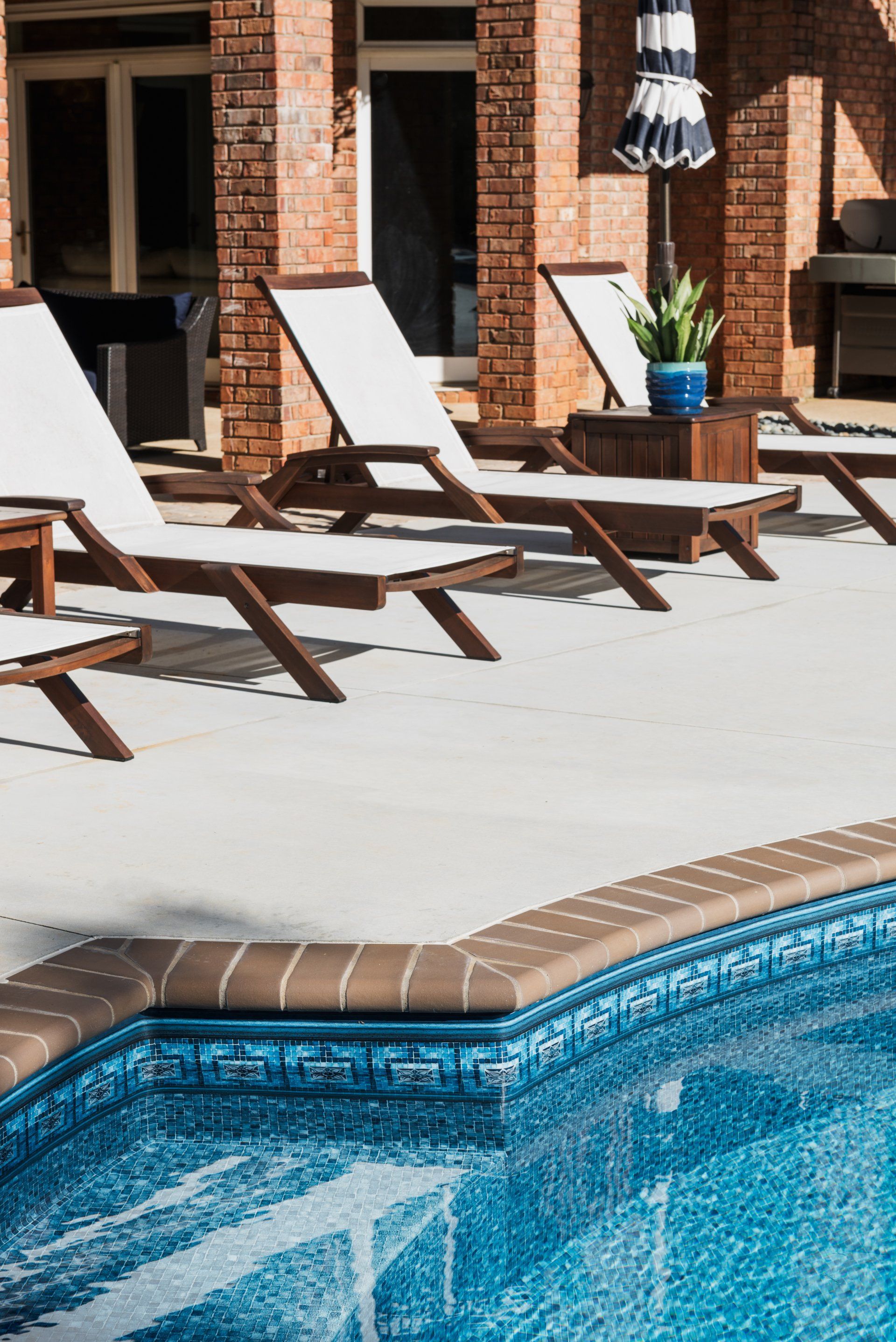 Poolside lounge chairs beside a blue swimming pool and brick patio with potted plants