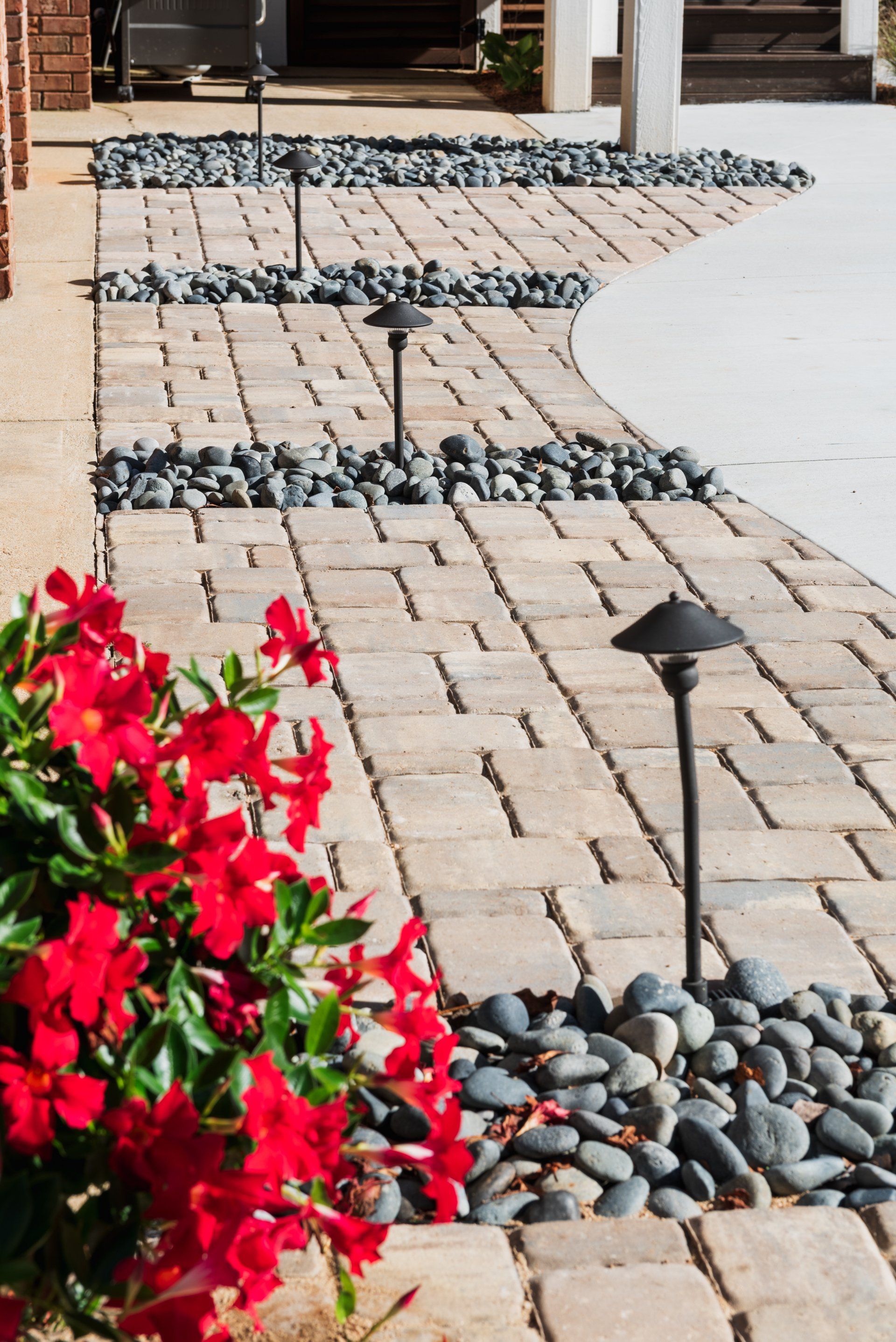 Brick walkway with dark decorative pebbles, black path lights, and bright red flowers beside a curved patio edge