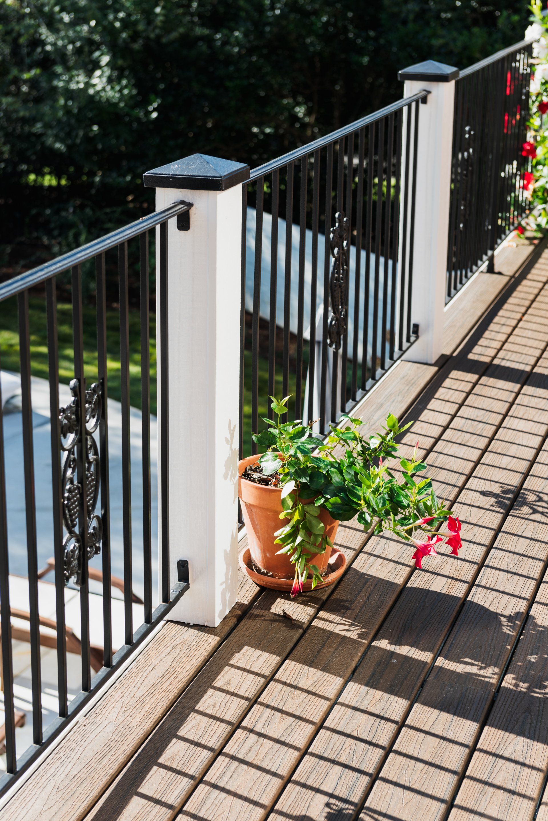 Balcony with black railing, sunlit wooden floor, and potted flowers beside a tiled wall