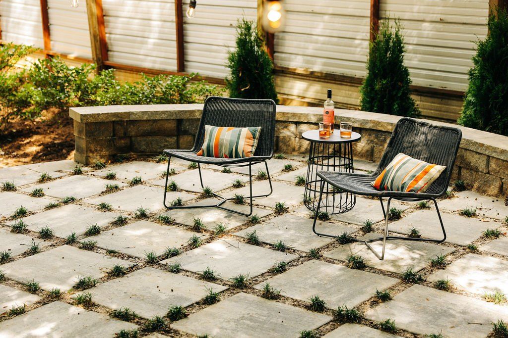 Two lounge chairs and a small table on a sunlit stone patio with potted plants and a brick wall