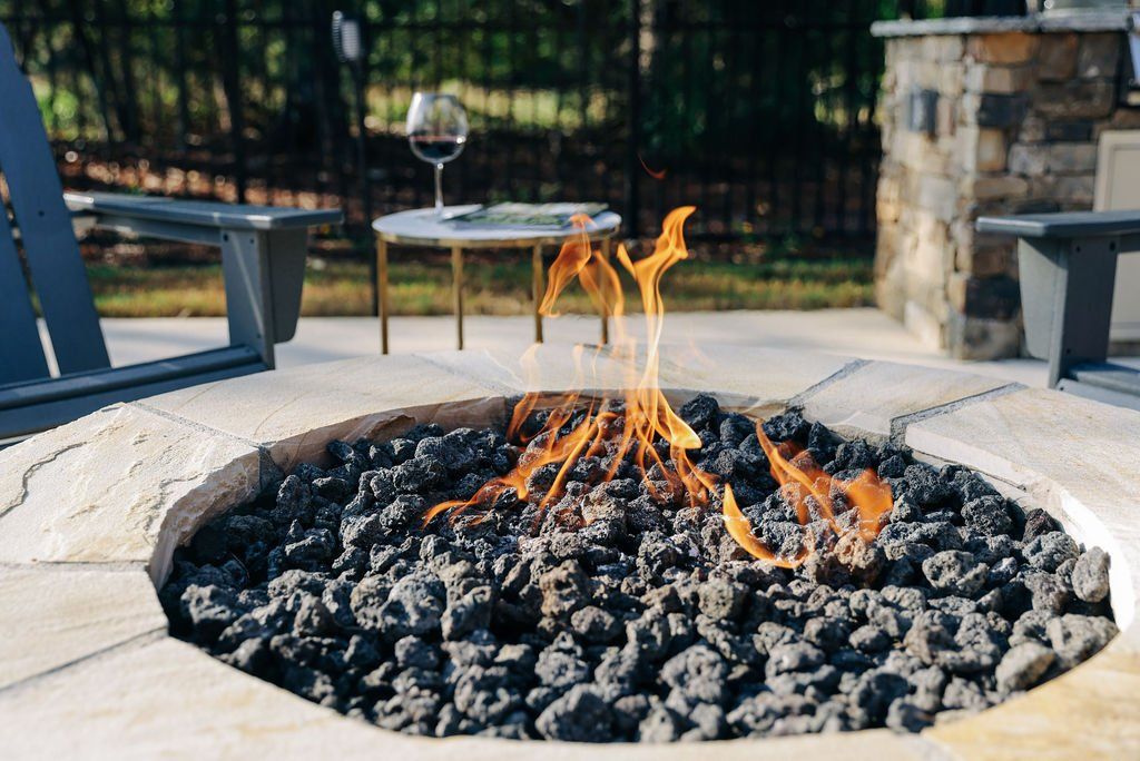 Backyard fire pit with flames over black stones beside patio chairs and a small table