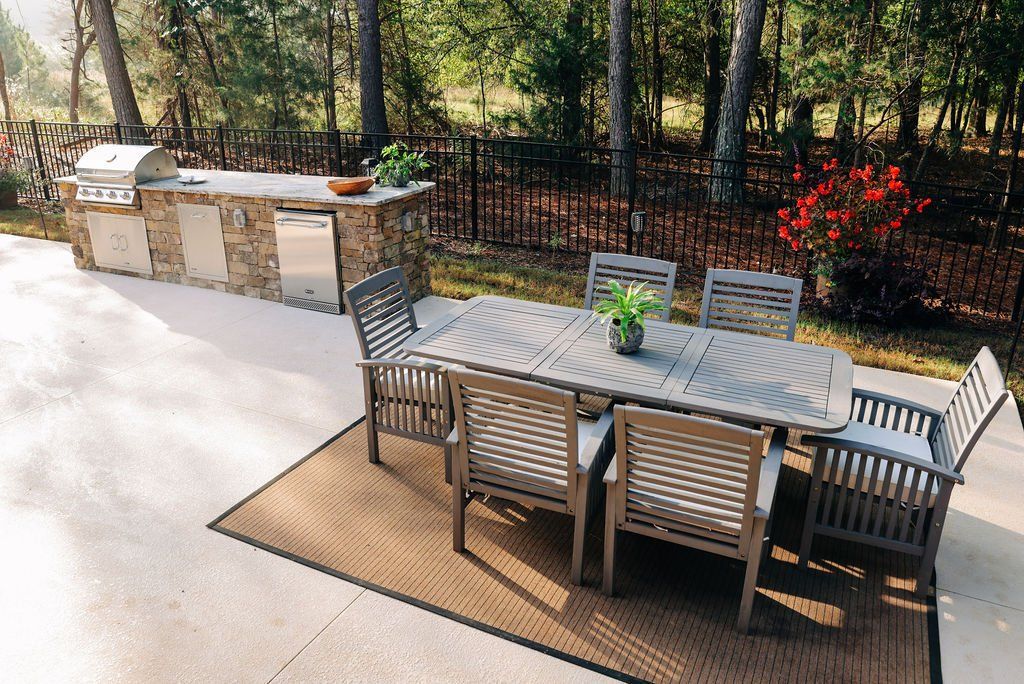Outdoor patio with a dining table, chairs, potted plants, and a stone grill area beside a fence.