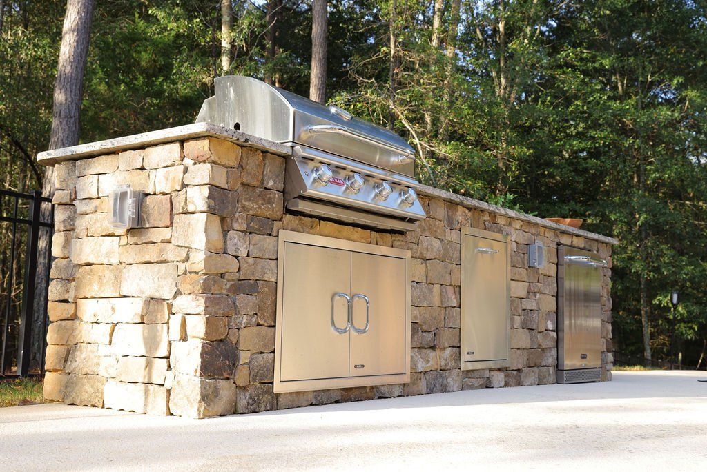 Stone outdoor kitchen with stainless steel grill and cabinets beside a wooded road