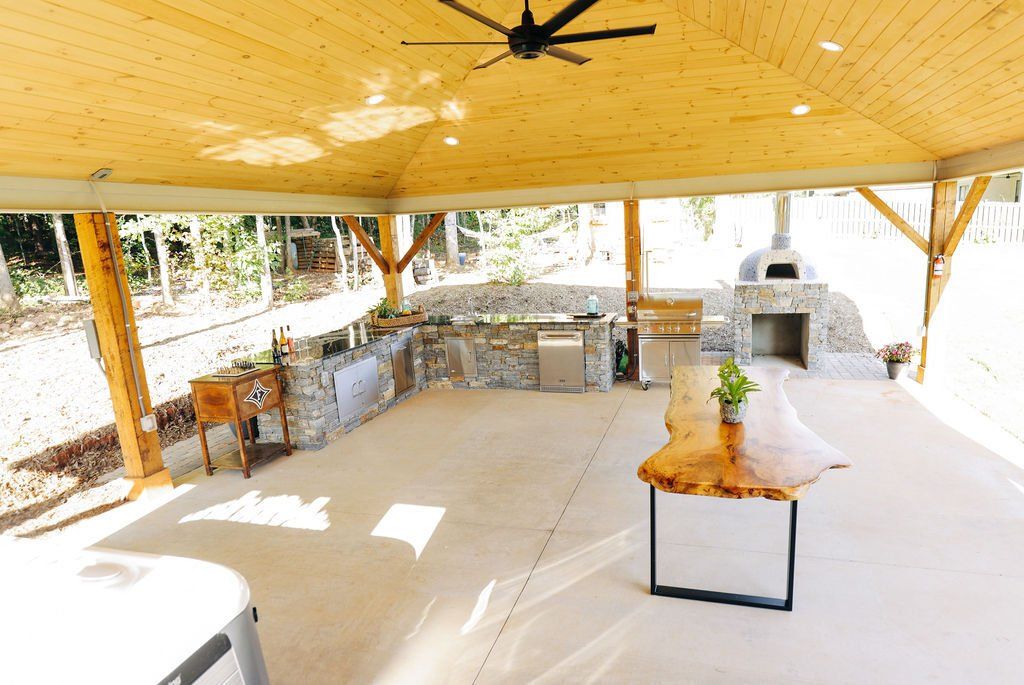 Open-air pavilion with stone counters, a wooden ceiling, and a serving table set with flowers and drinks.