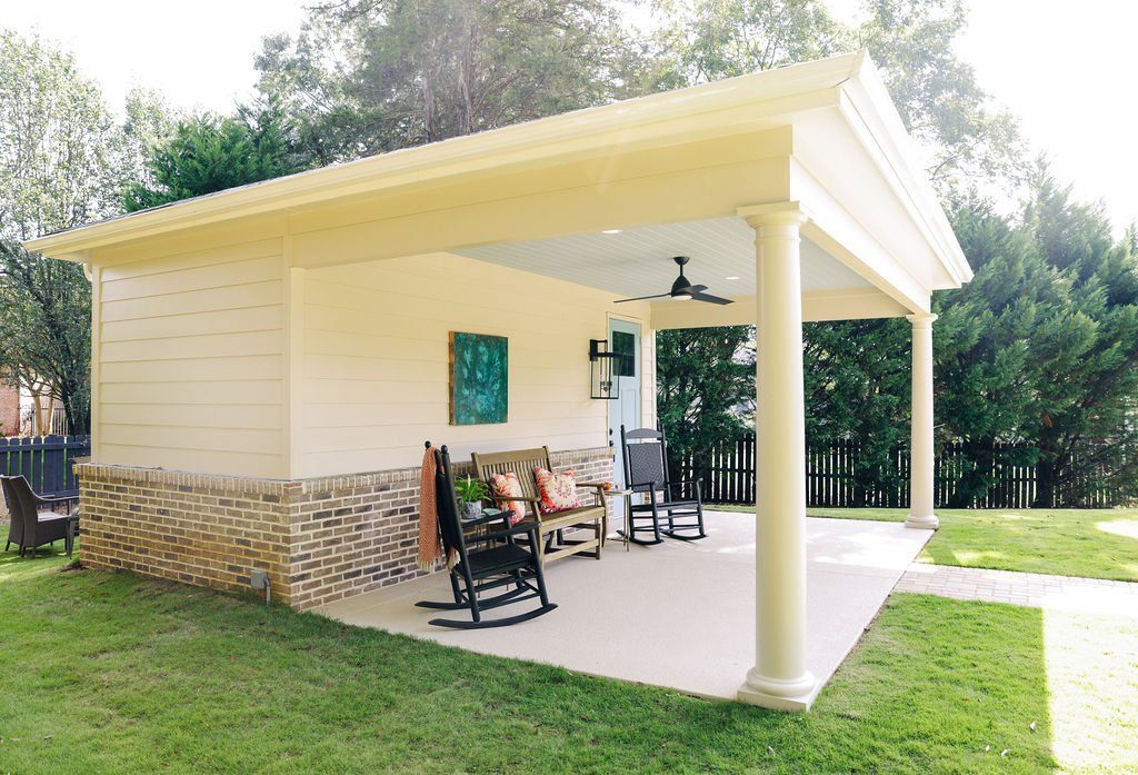 Small yellow house with stone base and covered porch, rockers, and white columns on a grassy lawn