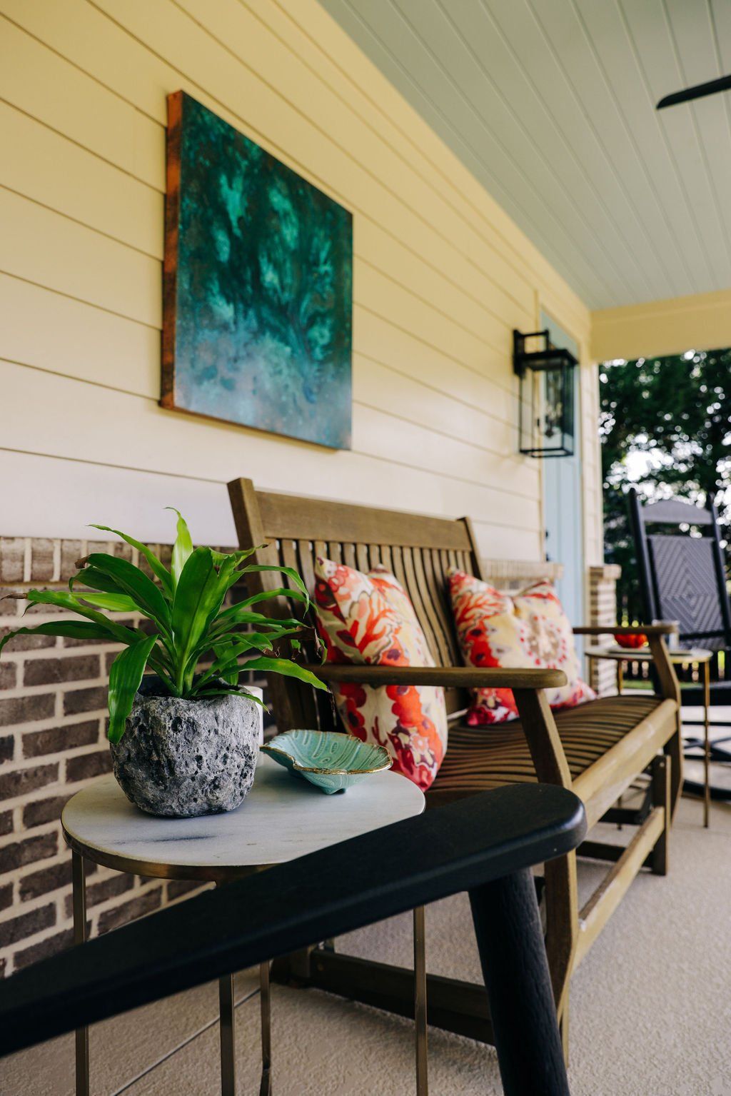Porch with wicker bench, floral pillows, small table with potted plant, and abstract wall art