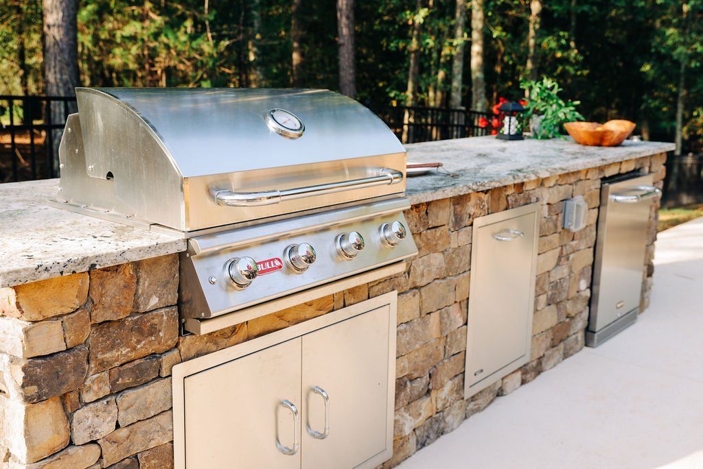 Outdoor stone patio kitchen with stainless steel grill, sink, and cabinets against a wooded backdrop