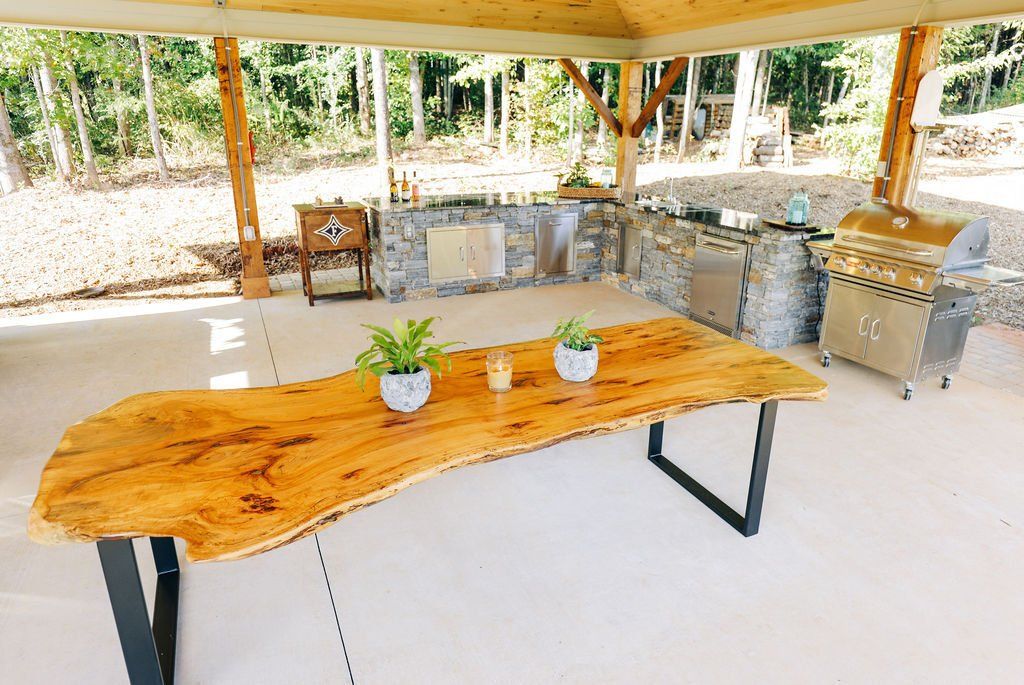 Open-air kitchen with a live-edge wooden table, stone counters, and grill under a pavilion.