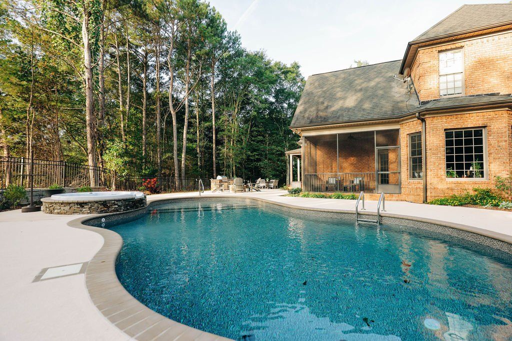 Curved backyard swimming pool beside a thatched-roof house and tall trees
