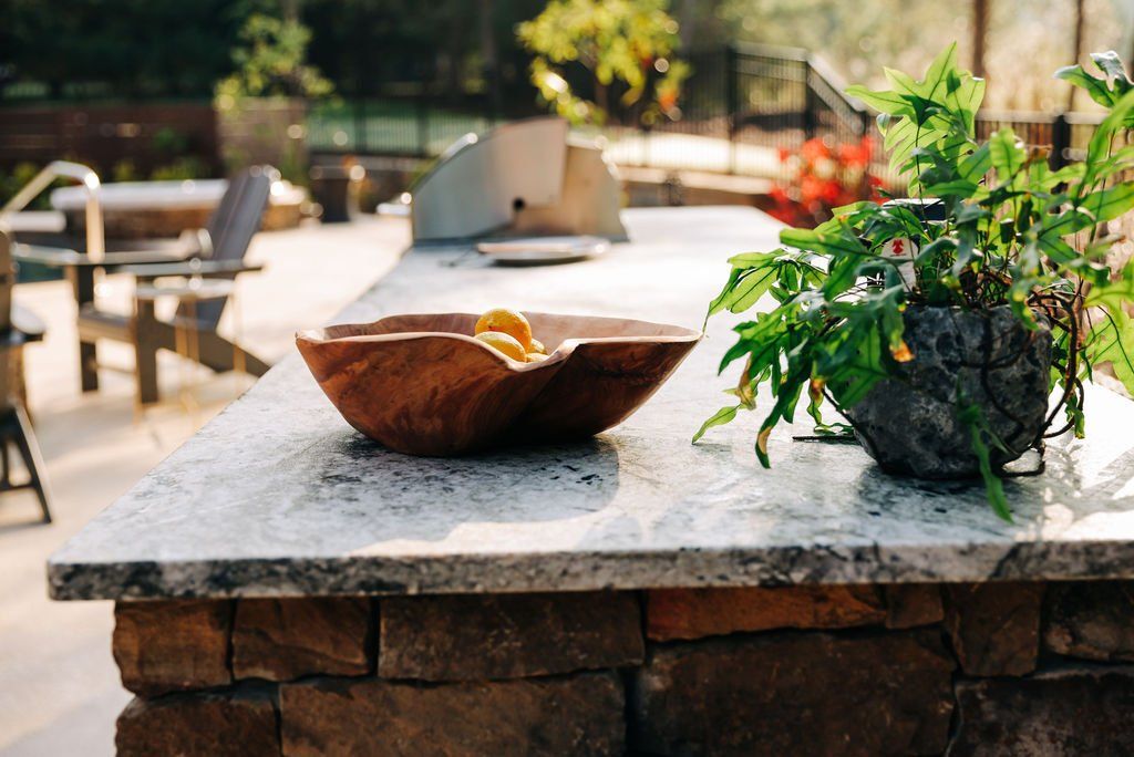 Stone patio table with wooden bowl and potted greenery in a sunny outdoor garden setting