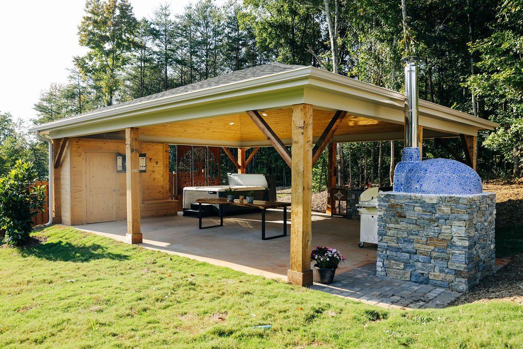 Open-air pavilion with wooden posts, picnic table, and stone fire pit in a grassy yard