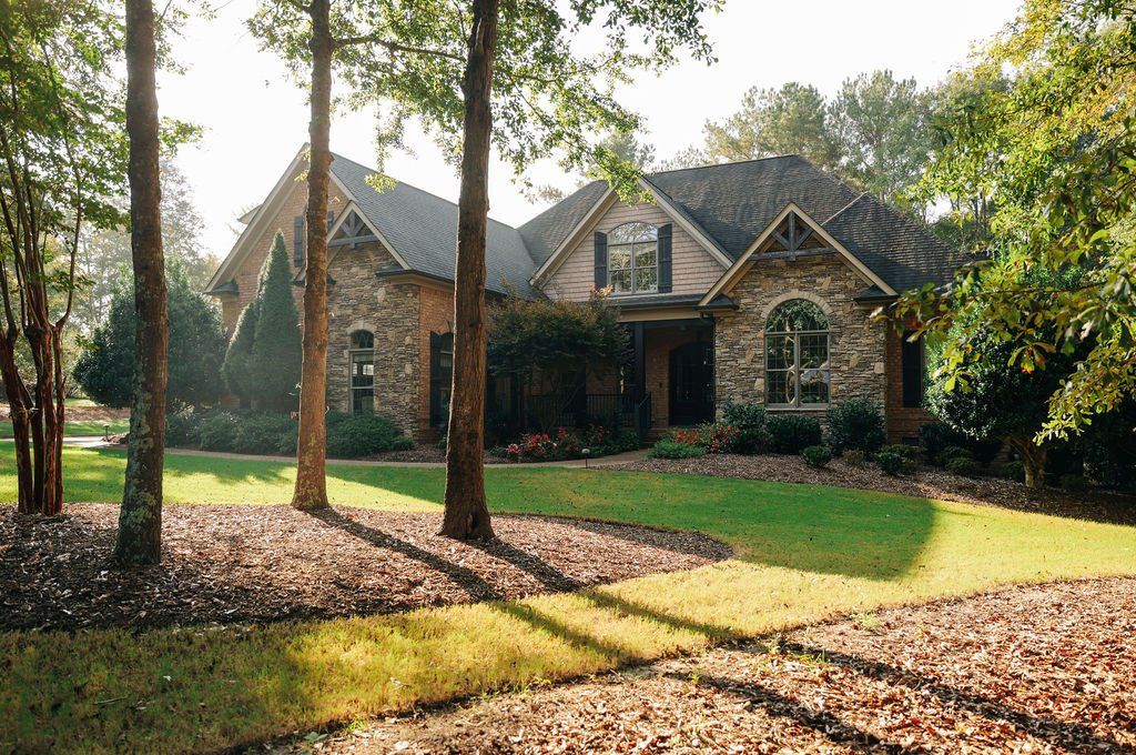 Brick house with gabled roof and arched windows, surrounded by tall trees and a sunlit front lawn.
