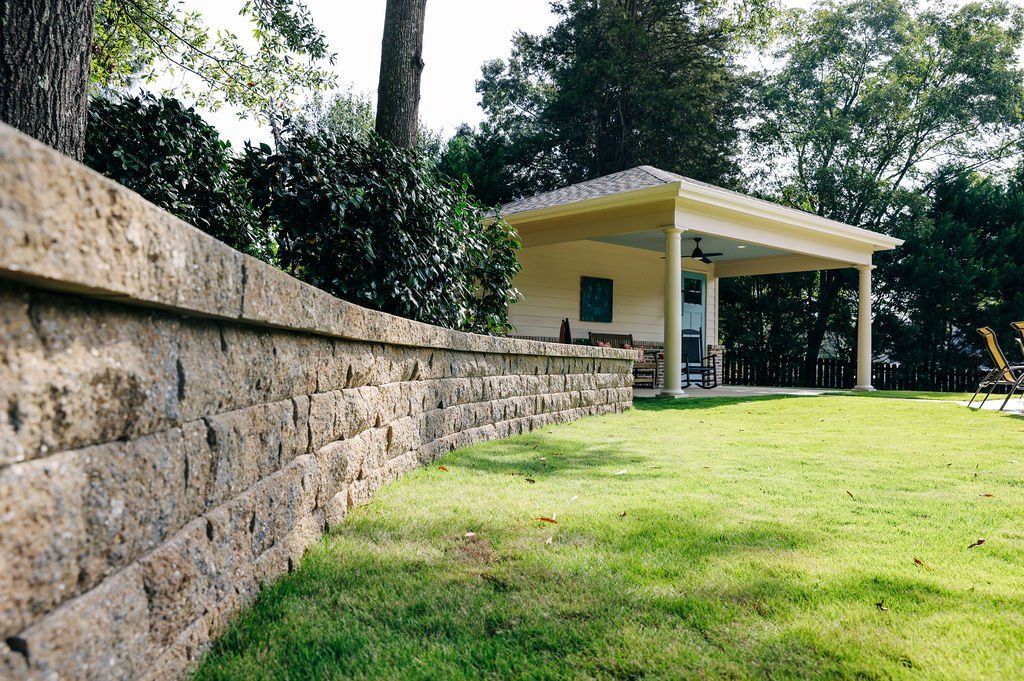 Stone retaining wall beside a grassy yard, with a small yellow pavilion under trees in the background