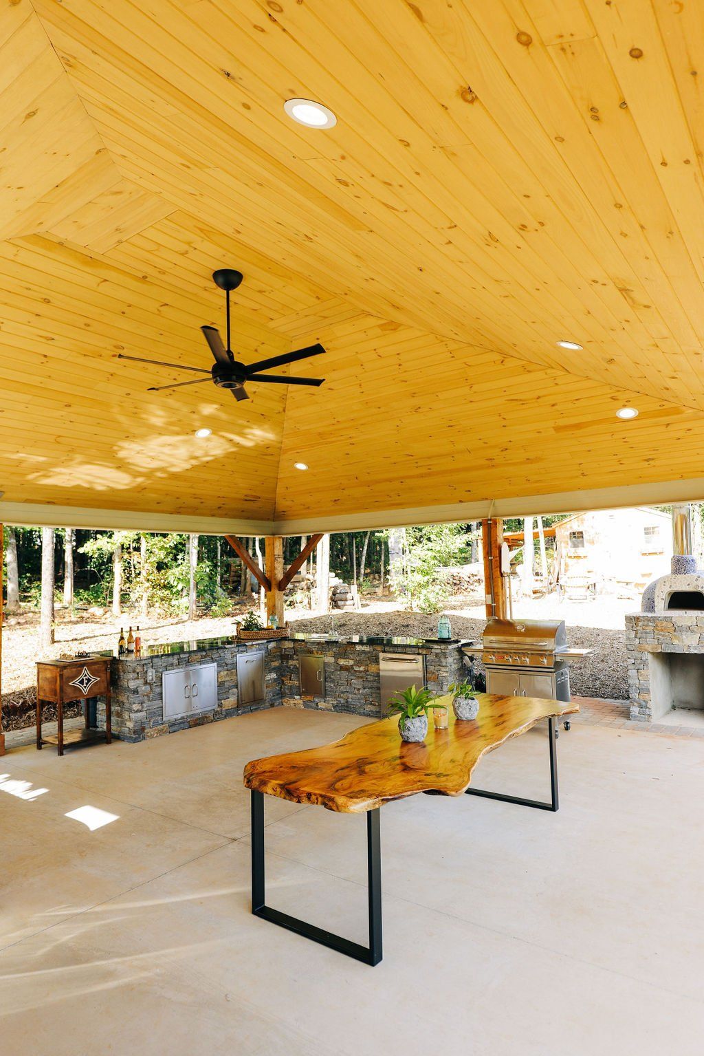 Open-air patio with wood ceiling, ceiling fan, stone counter, and a wooden table in the center