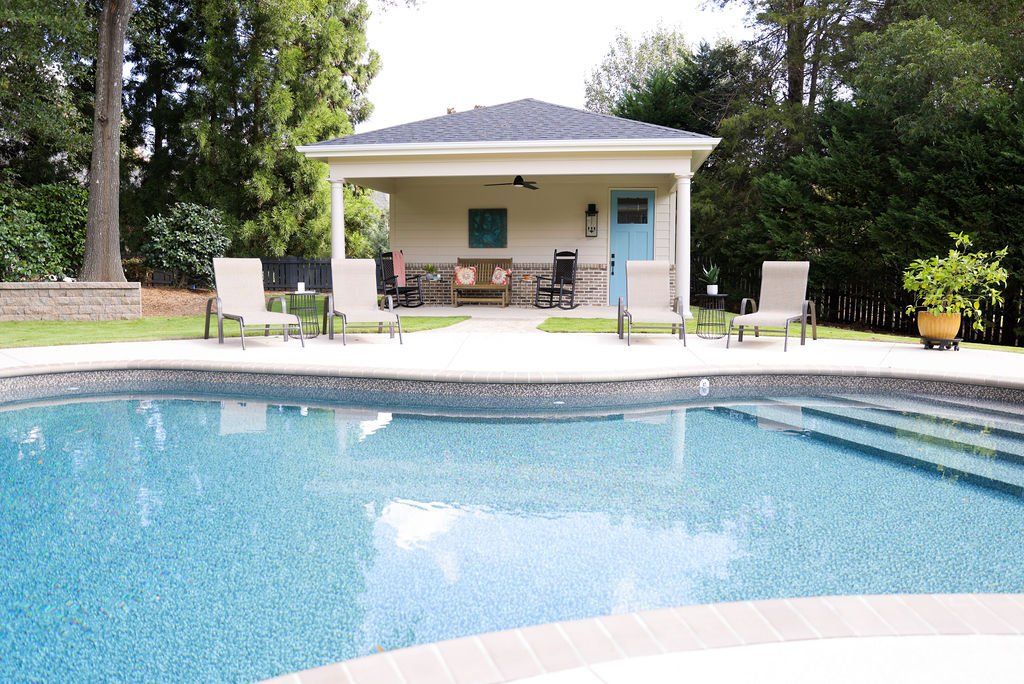 Backyard swimming pool with patio chairs and a small pool house surrounded by trees