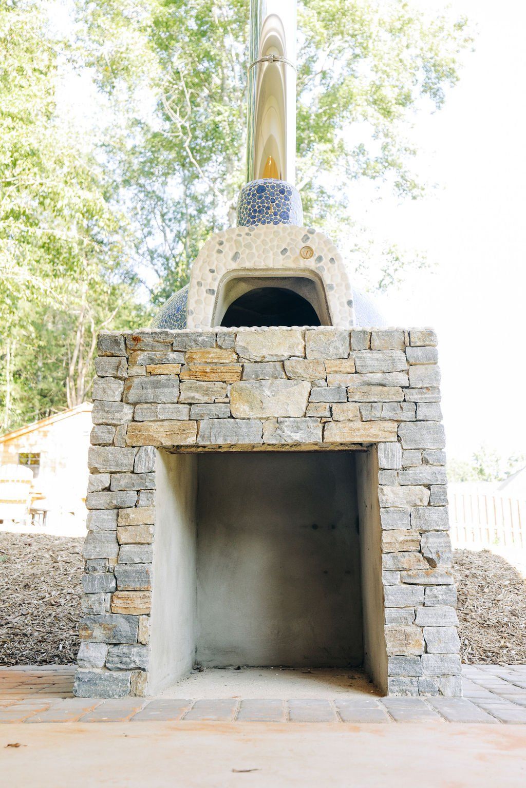Stone outdoor fireplace with a tall chimney and arched opening, surrounded by trees and sunlight