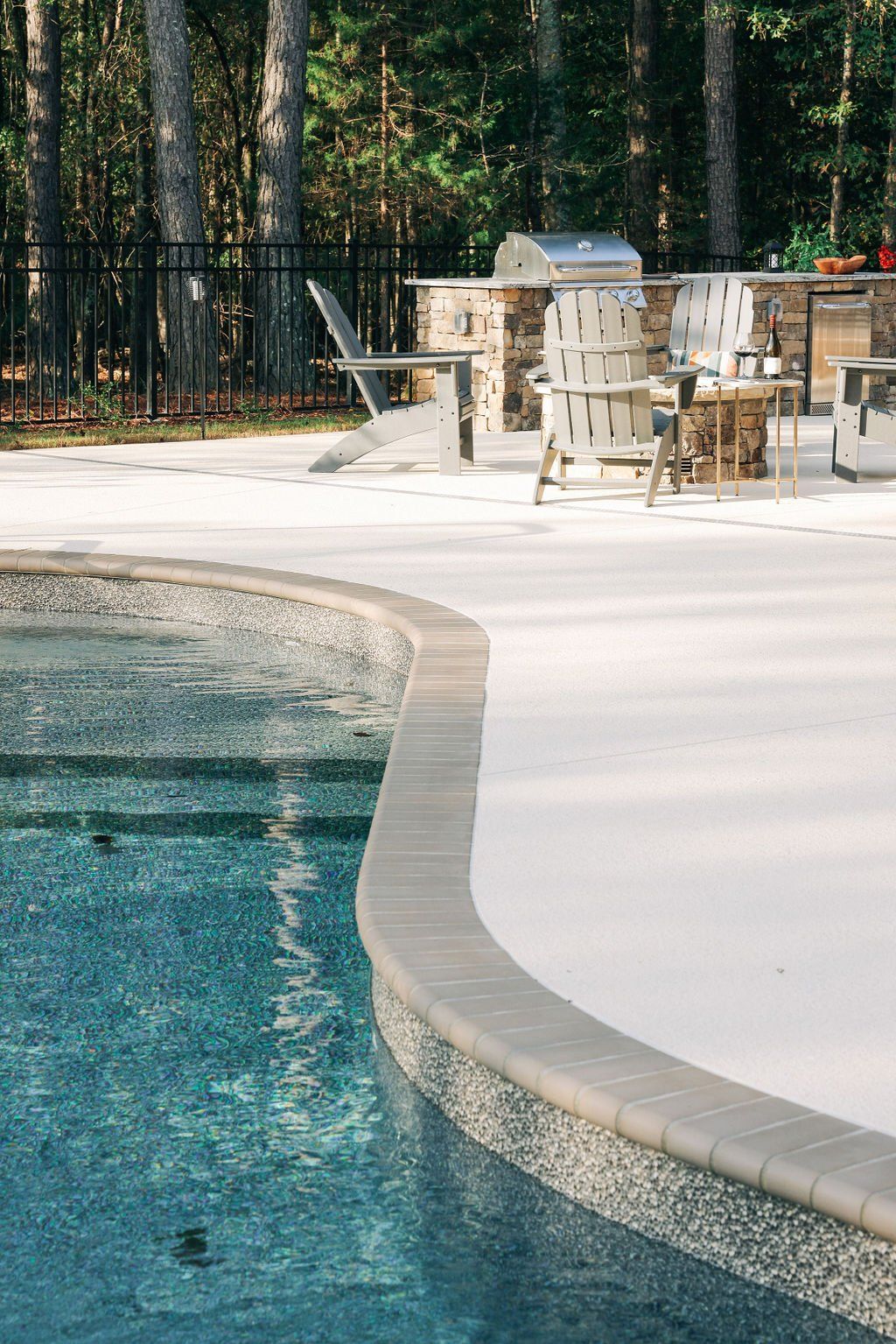Curved backyard swimming pool beside a patio with chairs and a grill, bordered by trees and a black fence
