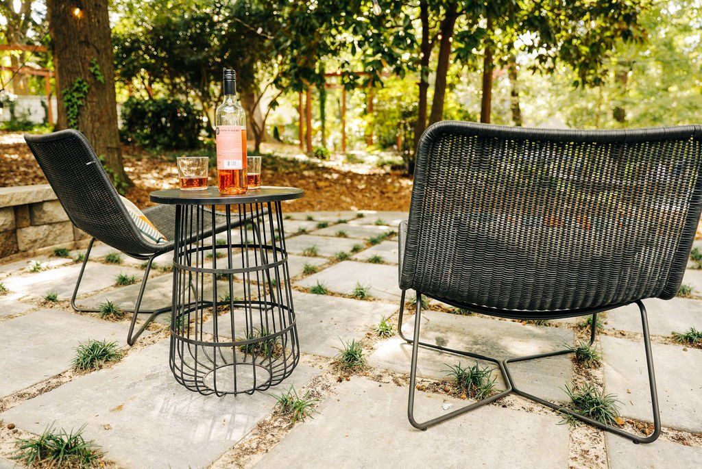 Outdoor patio with two black woven chairs and a small round table on stone paving in a wooded area