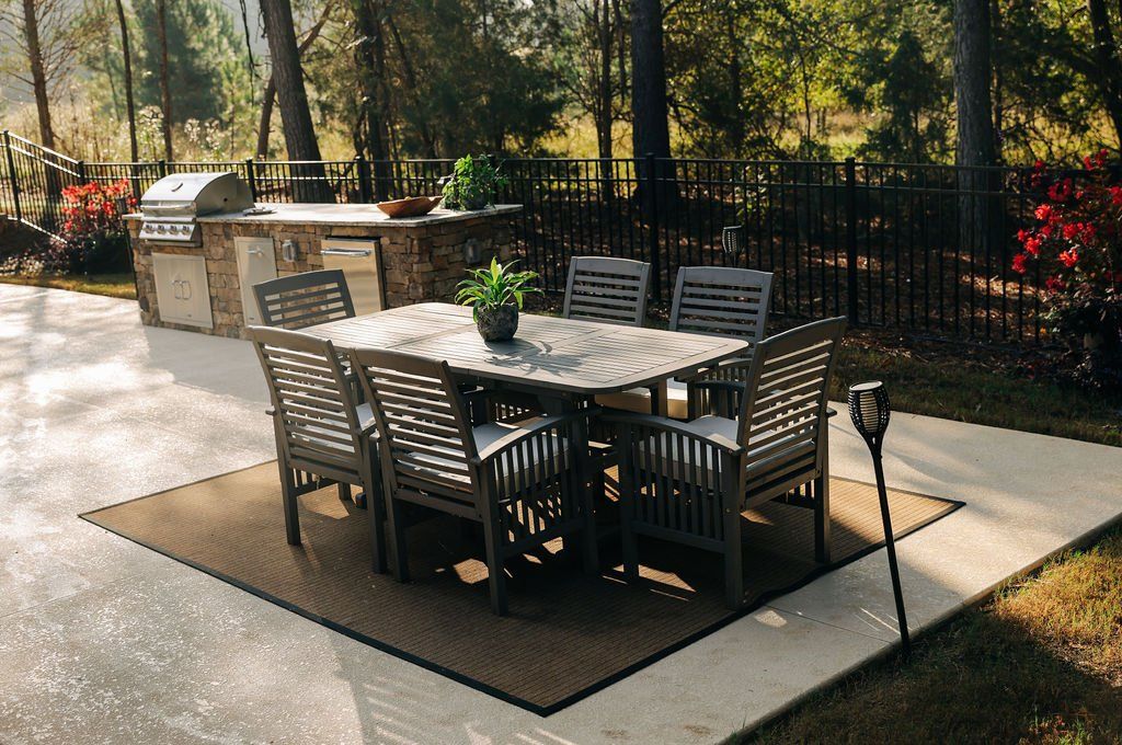 Outdoor patio dining area with a glass table, six chairs, grill, and garden fence in the background