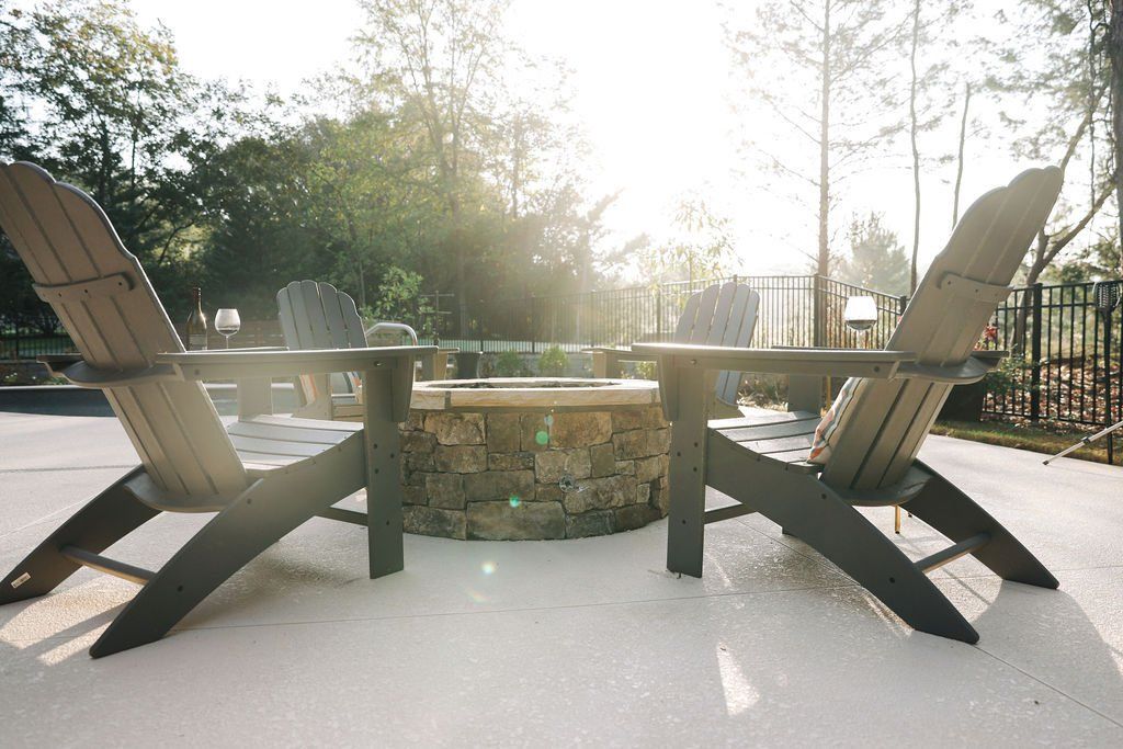 Two wooden Adirondack chairs around a stone fire pit on a sunny patio, with trees in the background