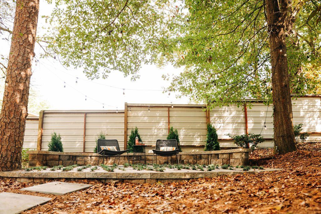 Leaf-covered yard with two large trees, a white fence, and potted plants along a stone border